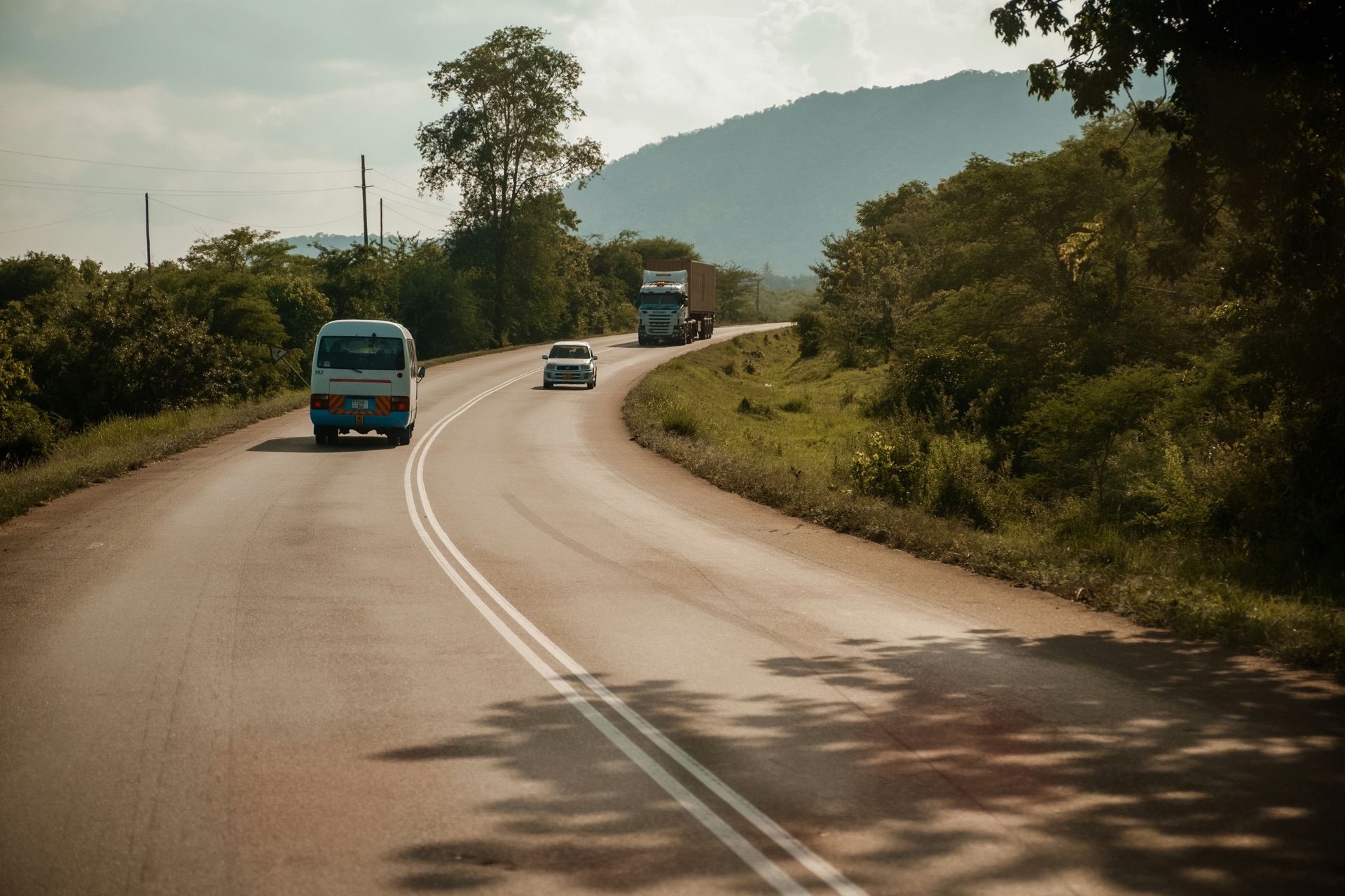 A two-lane paved highway curving through green hills, with a minibus, a car and a container truck.