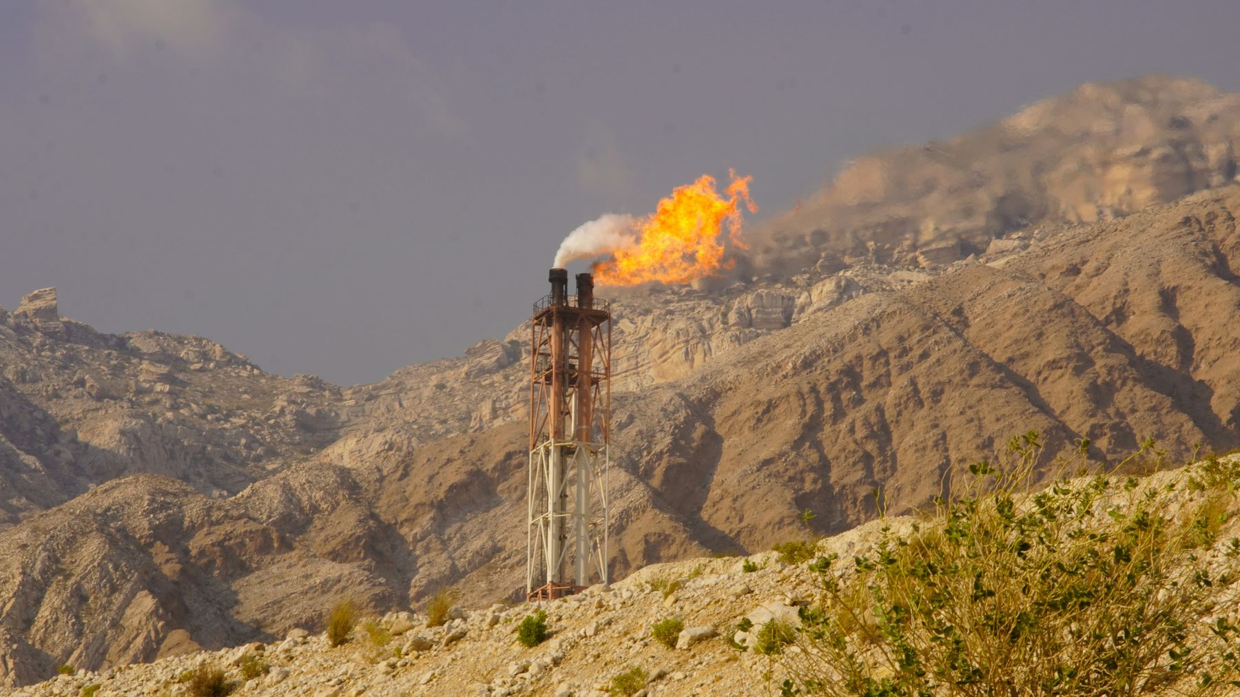 A gas flare burning at a wellhead stack against arid mountains.