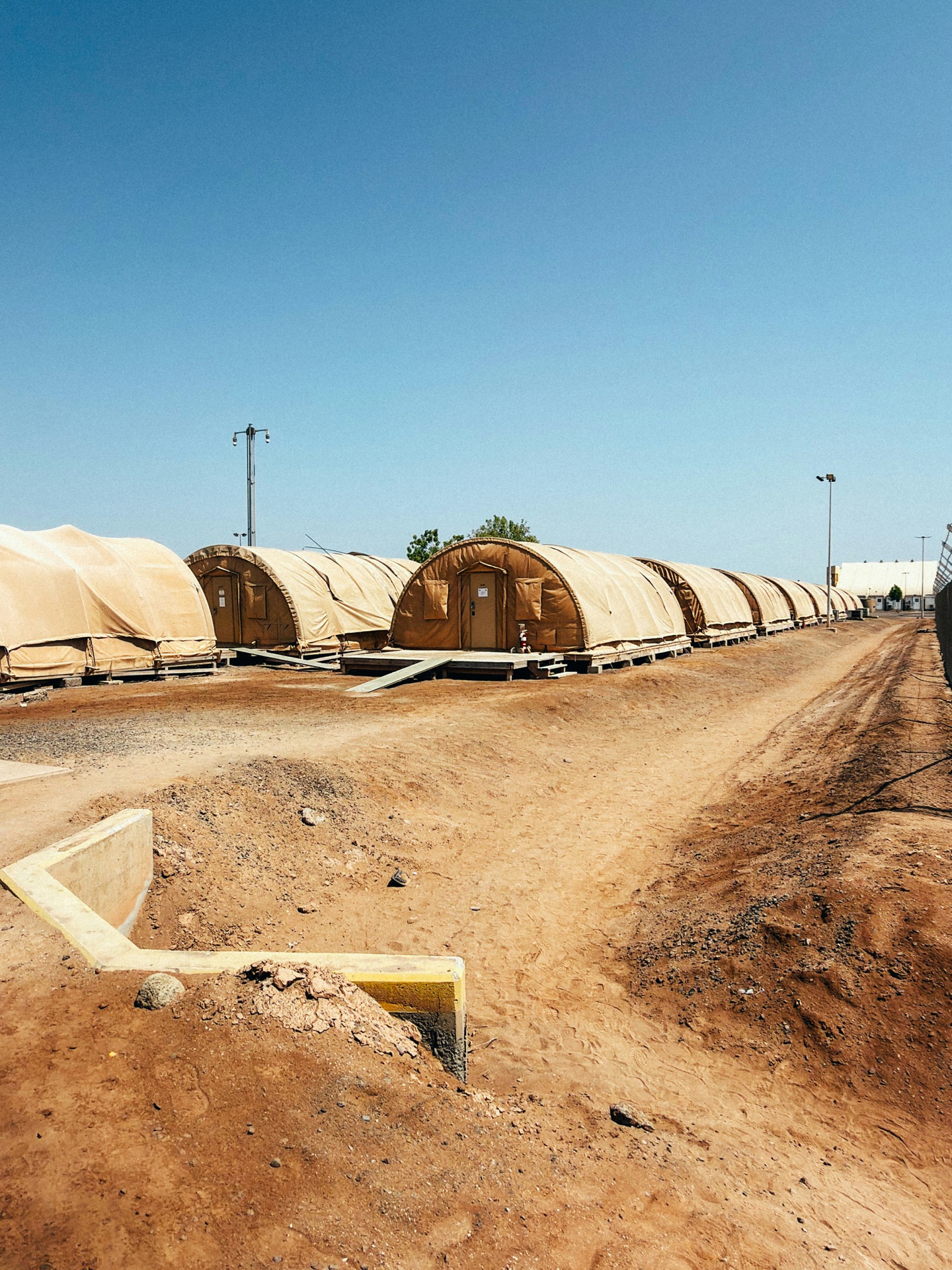 Rows of shelters on raised platforms beside an unsealed dirt track and a concrete drainage channel.
